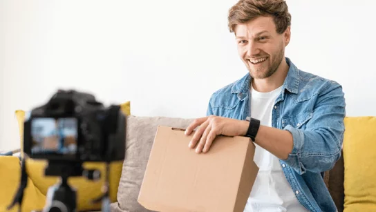 Lachender Mann mit einem Karton vor einer Kamera